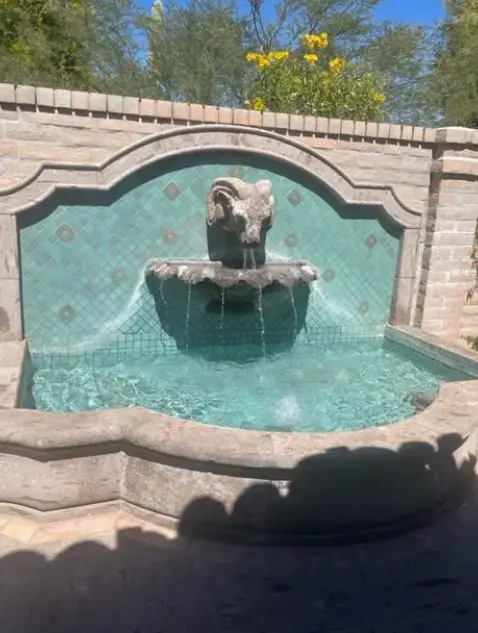 A decorative stone fountain featuring a ram's head, with turquoise tiles and flowing water, surrounded by lush greenery