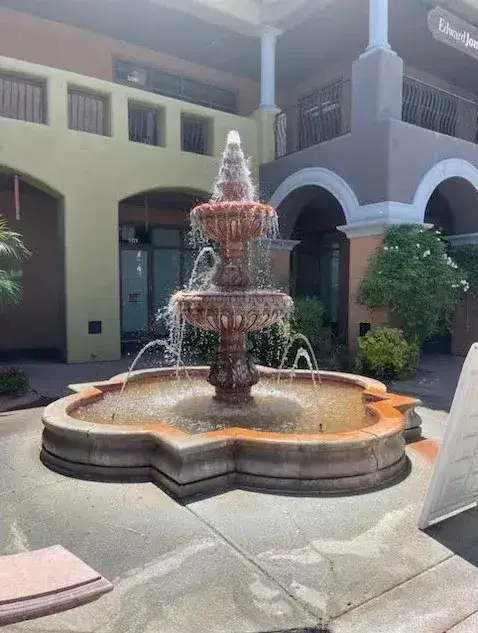 A three-tiered stone fountain with water flowing gracefully, surrounded by lush greenery and vibrant architecture