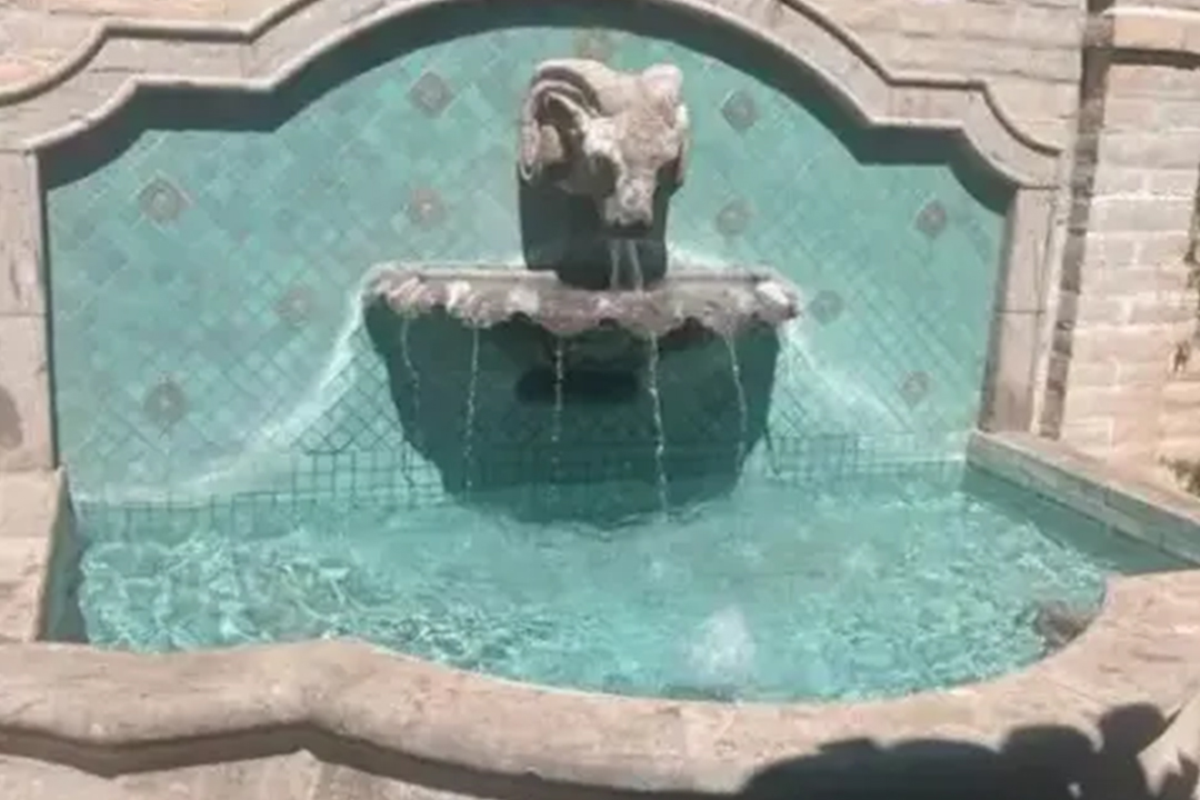 A decorative stone fountain featuring a ram's head, with water flowing into a turquoise-tiled basin under a sunny sky.