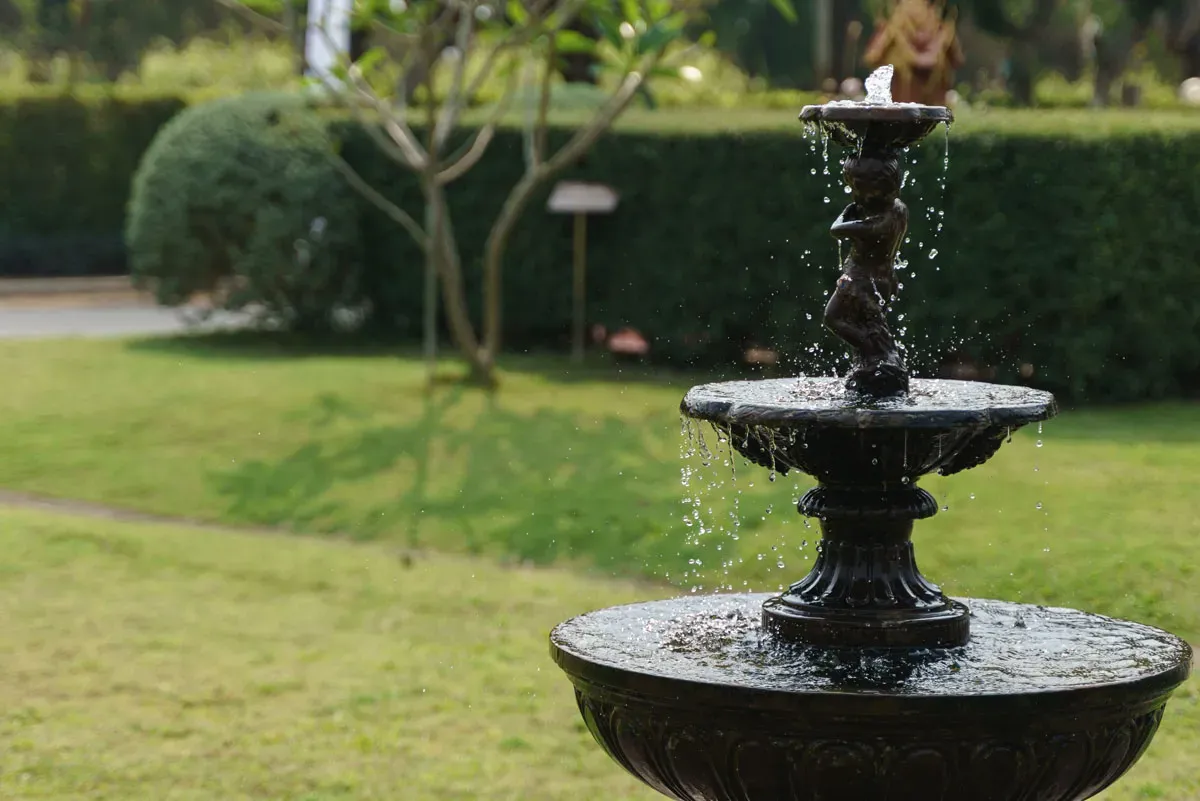 fountain-black.jpg A decorative fountain with flowing water, featuring a sculpted figure at the top, surrounded by a lush green garden