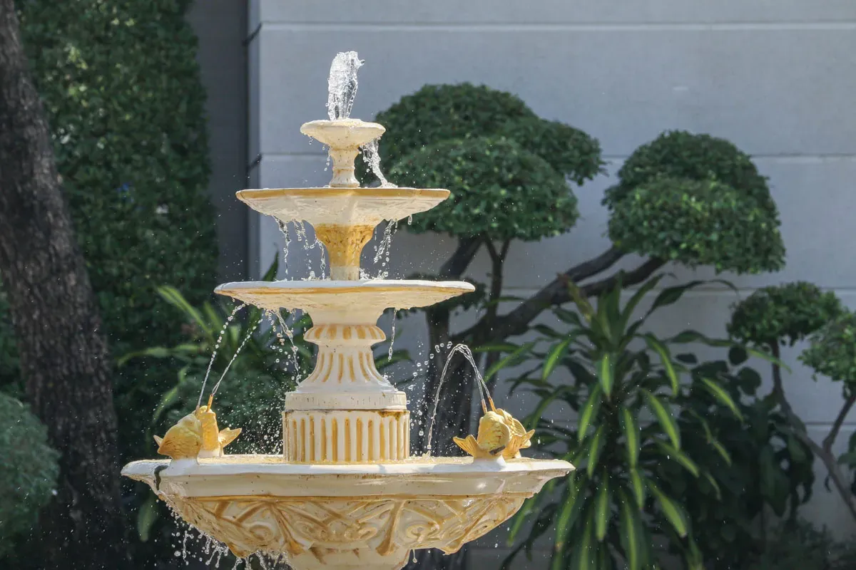 fountain-yellow.jpg A decorative three-tiered fountain with golden details, water spouting from the top and fish motifs, surrounded by lush greenery
