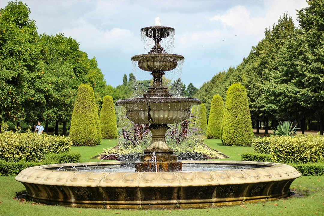 A three-tiered fountain surrounded by lush greenery and neatly trimmed hedges in a serene park setting under a blue sky.